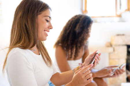 Two happy mixed race women friends having fun at home with social network chatting on smartphone with internet mobile connection technology. New normal human habits for mutual contacts between peopleの写真素材