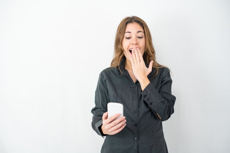 Studio shot of young model woman in grey shirt business clothes isolated on white copy space background surprised looking at smartphone with wow expression hand on mouth. Concept of new technologiesの写真素材