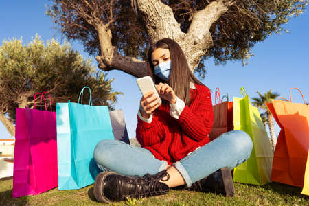 Teenager sitting under tree among colored shopping bags wearing Covid-19 medical protection mask...