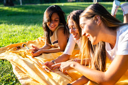 Three young happy women having fun with internet mobile technology using smartphone lying on meadow...