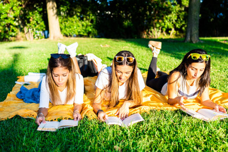 Three young women students spending time lying on meadow in city park at sunset or dawn reading book to learn university school lesson. Girl relaxing in city park enjoying preferred romance taleの写真素材