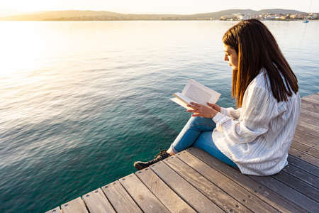 Young woman sitting on a pier at sunset or sunrise on transparent sea ocean water reading a book. Student girl learn university school lesson in nature. Bookaholic person having fun with romance taleの写真素材