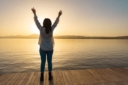 Silhouette of brunette girl view from back in transparent shirt standing on a pier with open arms up to the sky looking at horizon with setting sun with reflection on sea ocean water. Pensive conceptの写真素材