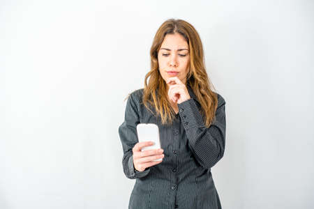 Studio shot of Caucasian pensive young woman holding chin watching smartphone wearing business clothes. Worried gesture by thoughtful person in stress using internet mobile technologyの写真素材