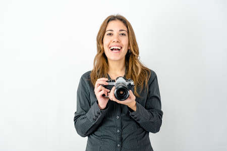 Studio shot of smiling happy young woman holding an old vintage photo camera watching photographer on white background for copy space. Concept of passion for imaging and history of classic photographyの写真素材