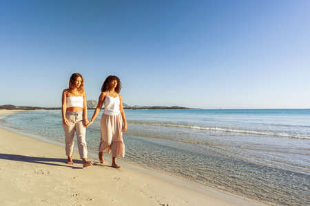 Multiracial couple of beautiful young gay women holding hands walking at sunrise or sunset on a tropical colored beach with flat ocean sea water. Concept: in love and nature there are no differencesの写真素材