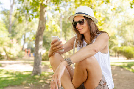 Young beautiful happy in light clothes enjoy her summer vacation in the city park using smartphone to take part in social network community sharing pictures and video of life moments of her travelの写真素材