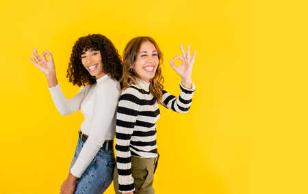 Two multiracial young beautiful models posing with ok gesture in toothy smile isolated on yellow background for copy space. Concept of positive mood, work success, and happiness enjoying your lifeの写真素材