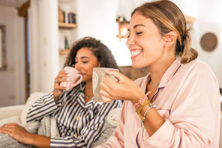 Couple of cohabiting girls in pajamas relaxing under blanket on cozy sofa drink a cup of tea while...