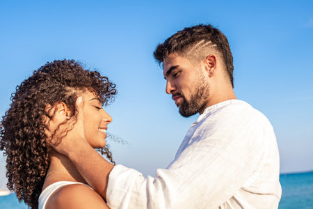Moment of tenderness between young multiracial couple of happy lovers who look into each other's eyes while he smilingly holds her head in hands. Passion and love of two Latin people on sea vacationsの写真素材