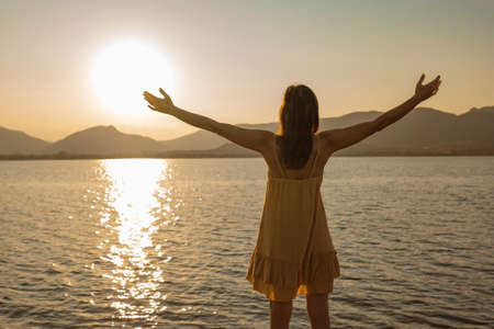 Lonely pensive woman with open arms in adoration of sun reflecting in ocean sea water at sunset or sunrise. Concept of spirituality and serenity in contact with nature to find yourself and one's soulの写真素材