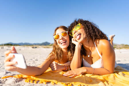 Two beautiful girls lying on the beach in the summer having fun making faces with their tongues out while taking a selfie. Two multiracial lesbian girlfriends with funny sunglasses using smartphoneの写真素材