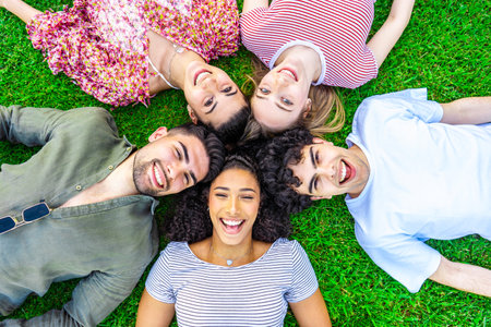 Group of five happy young diverse multiracial gen z friends lying in circle head to head on the grass of a park lawn looking to the camera from the bottom. Concept of friendship without prejudicesの写真素材