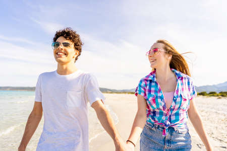 Carefree youth concept: young very happy gen z real couple holding hands tenderly walking on the seashore of tropical beach in summer with sunglasses. Girl in love looks intently at her boyfriendの写真素材