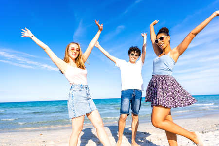 Three very happy friends having fun at the beach posing laughing looking at camera for a portrait with raised arms. Gen z multiracial carefree people in tropical beach with white sand and blue skyの写真素材