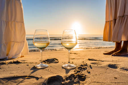 Sunset or sunrise beach party scene with two unrecognizable multiracial boho girls wearing long white dress barefoot on the ocean beach sand. Wine glasses on the ground with sun reflections effectの写真素材