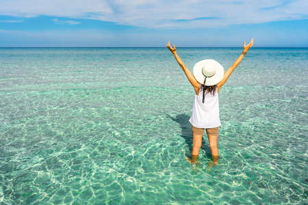 View from back of hipster unrecognizable millennial happy young woman with a large white hat in vacation resort on a crystal clear tropical sea water open arms up to the blue sky looking to horizonの写真素材