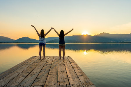 Two girls focused on spiritual meditation looking at setting sun standing on a wooden pier on flat water with light reflections. Suggestive sunset scene of pensive young women in contact with natureの写真素材