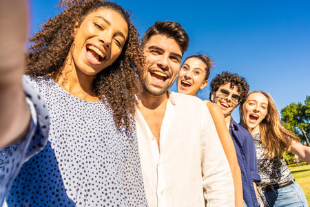 Five young multiracial friends in line posing for photo portrait smiling with open mouth. Black Hispanic brunette girl taking a selfie with smartphone outdoor. POV of happy millennial carefree peopleの写真素材