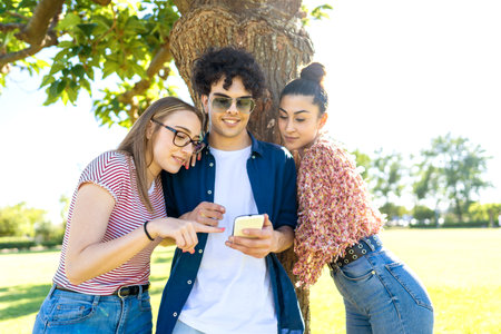 Three students having fun using smartphone at park pointing and texting on display. Two beautiful girls close to their best guy friend sharing on social network happy vacations momentsの写真素材
