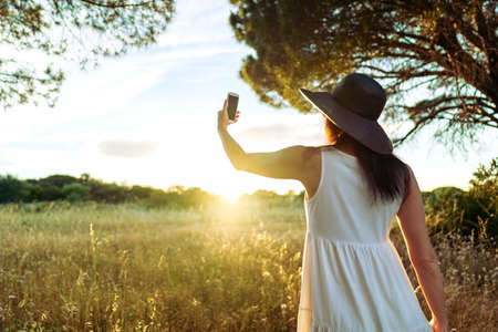 Suggestive scene from back of unrecognizable woman in white boho style dress and large dark hat taking a self portrait in front of the sun setting in the nature. Girl using cell for a self portraitの写真素材