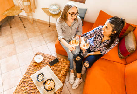 Couple of young women friends drinking at home sitting at orange sofa celebrating with champagne or white wine. Girls using alcohol to having fun taking aperitif in living room instead of study lessonの写真素材