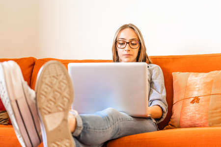 Cute blonde girl with eyeglasses sitting at home sofa focused on study or work at laptop computer with feet resting on table. Young self entrepreneur woman working from home using notebook and wifiの写真素材