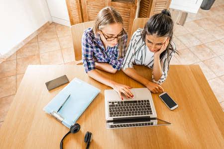 Two diverse girls view from top working or studying at laptop computer sitting at home table with note book, pen, headphone and smartphone. Gen z students using wifi to Attend online classes togetherの写真素材