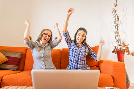 Two very happy girl cheering with hands up looking at laptop screen sitting at home sofa. Young winner women having success in their work. Change your life and make preferred job to live betterの写真素材