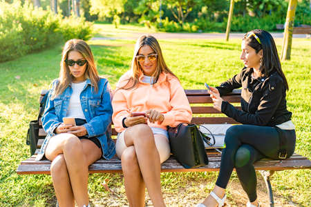 Three young woman best friends sitting on a city park bench at sunset or dawn using telephone to...