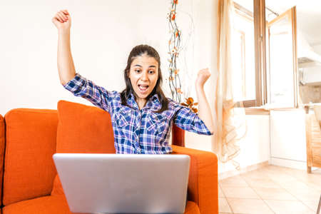 Brunette smart gen z girl rejoices in front of with open raised arms sitting at home sofa. Shocked young woman looking at computer screen in living room. Happy student looks results of a school examの写真素材