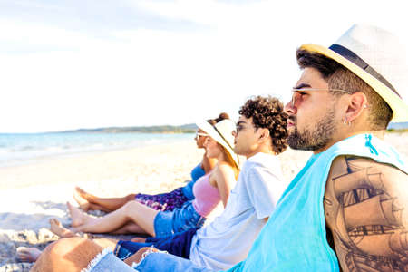 Selective focus portrait of handsome hipster guy with tattooed arm in white Panama hat sitting on seashore with friends looking at ocean water in vacation resort. Modern young people enjoying travelの写真素材