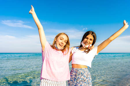 Two happy gen z girls embracing to each other with raised open arms looking at camera wearing funny protective open face mask enjoying sea vacations with crystal sea water and blue sky in backgroundの写真素材