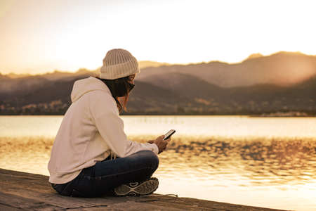 Unrecognizable young woman in white wool hat sitting on a pier alone wearing protective face mask using smartphone to keep in touch with friends and family due to social distancingの写真素材