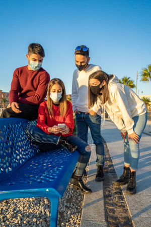 Millenial friends using smart phones wearing protective face mask on wave. Worried guys and girls watching news on mobile smartphone. Travellers group stay in touch during pandemic in vacationsの写真素材