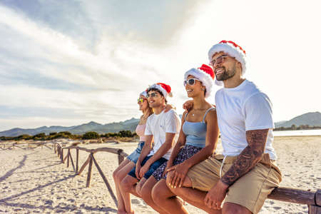 Perspective wide angle view of tattooed beautiful guy embracing his Hispanic girlfriends sitting on a wooden fence on a tropical beach enjoying holidays vacation sunglasses and Santa claus hatの写真素材