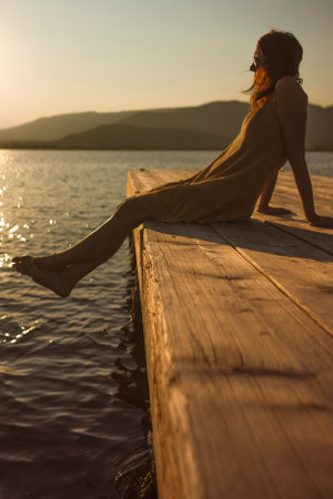 A contemplative young Caucasian woman sits alone on a lakeside pier, gazing at the water at sunset or dawn, in orange and vintage atmosphere.の写真素材