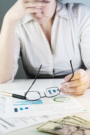Business woman analysing financial charts, supporting her head with arm and holding eyeglasses. Visible pen and dollar banknotes on white desk. Accounting, business, finance, tax and office concepts.の写真素材