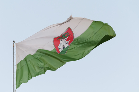 Closeup of weathered and torn flag of the city of Ljubljana waving in the wind. White-green flag with red emblem on the flagpole with bluish sky background. Capital of Slovenia.の写真素材