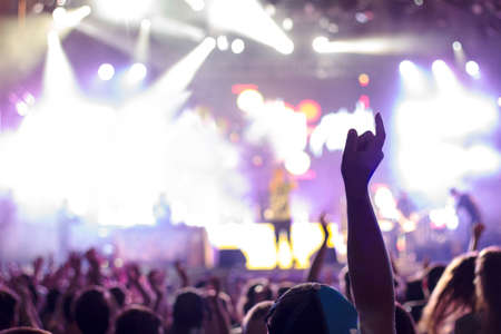 Night concert crowd with one hand pointing to the sky. The stage in the background is out of focus and has multiple white lights and flaresの写真素材