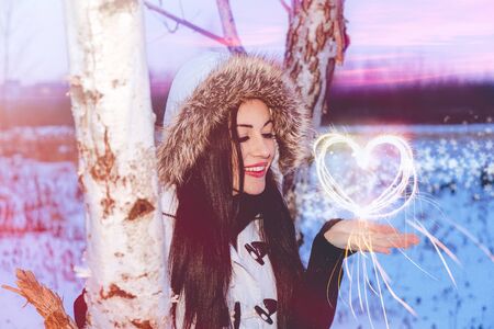 Snowing winter portrait of young woman brunette, close tree , pink lips , cathy brown eyes smiles hearts on handの写真素材