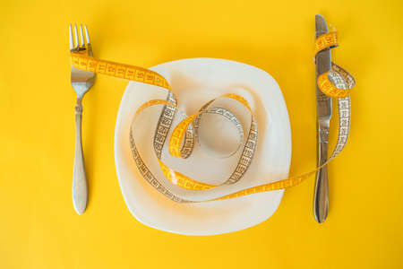 measuring tape on fork and plate and knife on a yellow background. body weight loss concept.の写真素材