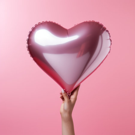 Female hand holding a pink heart shaped balloon on a pink background.の素材