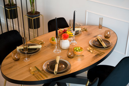 Elegantly set wooden oval dining table in a modern dining room. The table is prepared for a festive dinner party with dark plates, gold cutlery, and stylish glassware. The decor includes black candles, a vase with an autumn bouquet, and bowls with fresh grapes. This beautiful table arrangement is perfect for a special occasion, celebration, or holiday meal. Interior design concept.の写真素材