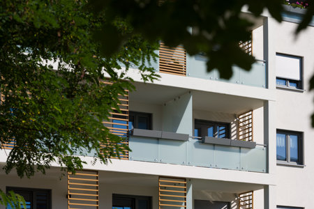 Exterior of a modern apartment building on a sunny day. The facade features contemporary balconies with frosted glass railings and decorative wooden slats. A green tree in the foreground adds a touch of nature to the urban setting. Concept for modern architecture, real estate, and city living.の写真素材