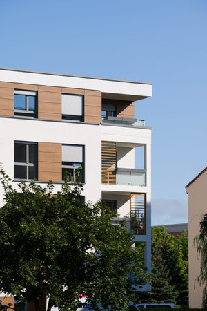 Exterior corner of a modern apartment building, featuring a white facade, decorative wooden panels, and multiple balconies. The balconies have glass railings and stylish wooden slats. The building is surrounded by green trees under a clear blue sky on a sunny day. This image represents contemporary architecture, real estate investment, and urban living.の写真素材
