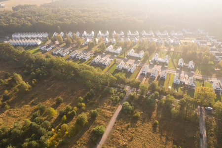 Aerial drone view of a modern housing estate with identical white houses located next to a green forest. The residential neighborhood is bathed in the warm, hazy light of a sunrise or sunset, creating long shadows. This image represents concepts such as real estate, property development, suburban living, and community.の写真素材