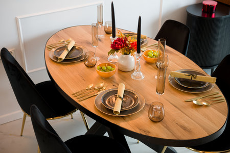 Elegantly set wooden oval dining table in a modern dining room. The table is prepared for a festive dinner party with dark plates, gold cutlery, and stylish glassware. The decor includes black candles, a vase with an autumn bouquet, and bowls with fresh grapes. This beautiful table arrangement is perfect for a special occasion, celebration, or holiday meal. Interior design concept.の写真素材