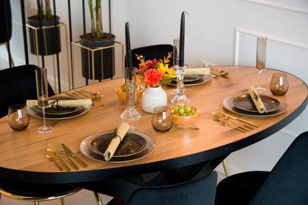Elegantly set wooden oval dining table in a modern dining room. The table is prepared for a festive dinner party with dark plates, gold cutlery, and stylish glassware. The decor includes black candles, a vase with an autumn bouquet, and bowls with fresh grapes. This beautiful table arrangement is perfect for a special occasion, celebration, or holiday meal. Interior design concept.の写真素材