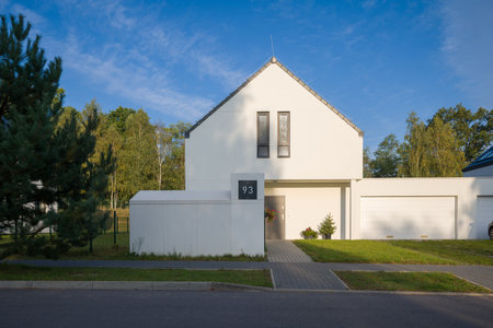 Exterior of a modern single-family house with a minimalist white facade and an attached garage. The property is located in a quiet suburban neighborhood on a sunny day. The front yard is landscaped with bushes, and the entrance is decorated for autumn. This image represents contemporary architecture, real estate investment, and suburban family living.の写真素材